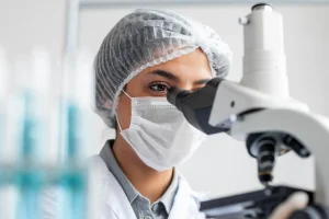 Cientista observando através de um microscópio em um laboratório, usando máscara e touca, cercada por frascos de vidro.