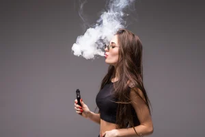 Mulher soltando vapor de um vaporizador, com longos cabelos e óculos, em fundo cinza. Ideal para temas de vaping e estilo de vida contemporâneo.